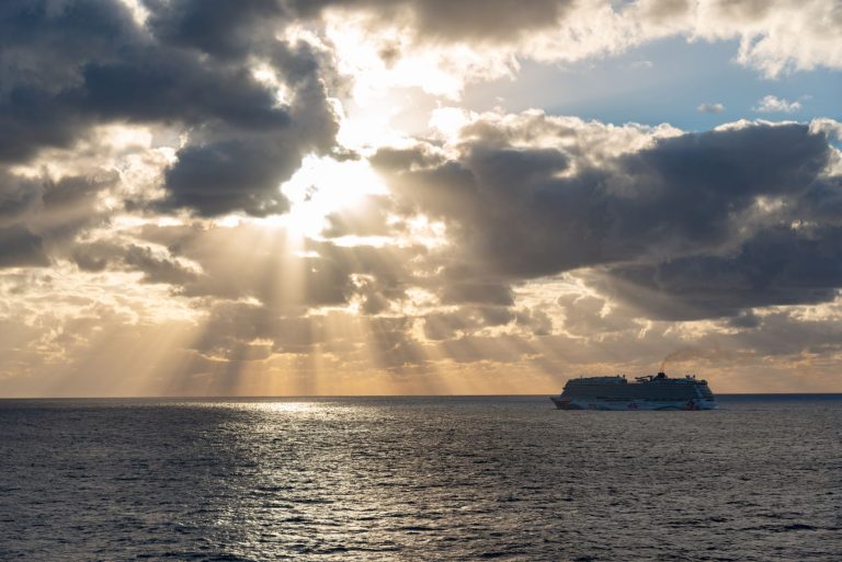 Navio de cruzeiro navega ao longe em um mar tranquilo sob a luz do amanhecer.